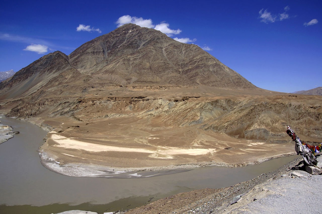 La montagne rocheuse de couleur beige semble descendre jusqu'à former une plage dans un méandre de la rivière Zanskar en direction de Chilling Ladakh